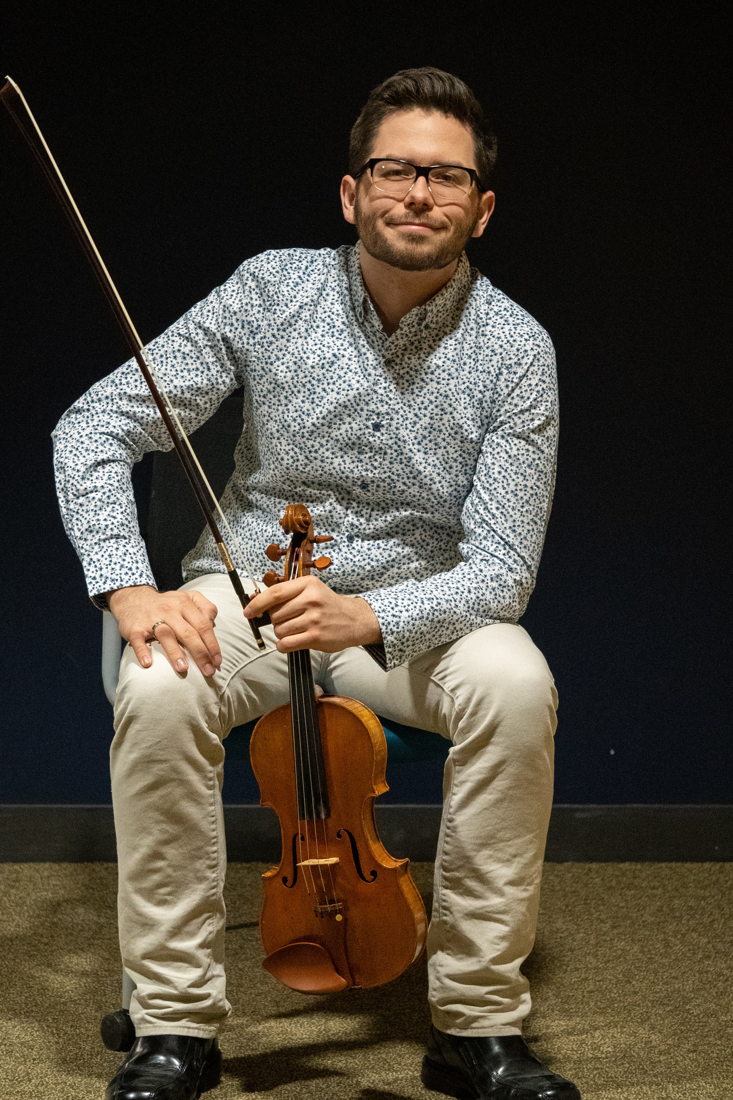 Mike is sitting in a chair, holding his violin and smiling at the camera